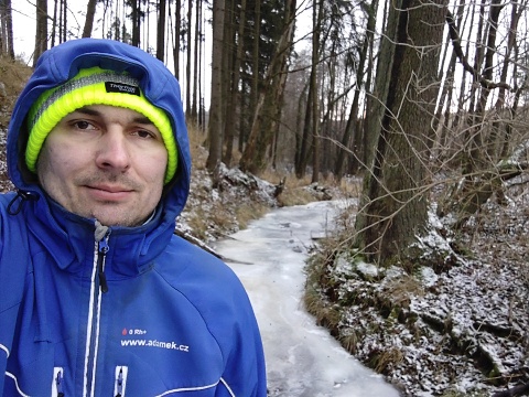 Martin Adámek – selfie w przyrodzie, niebieska kurtka, żółta zimowa czapka, na tle zamarznięty potok